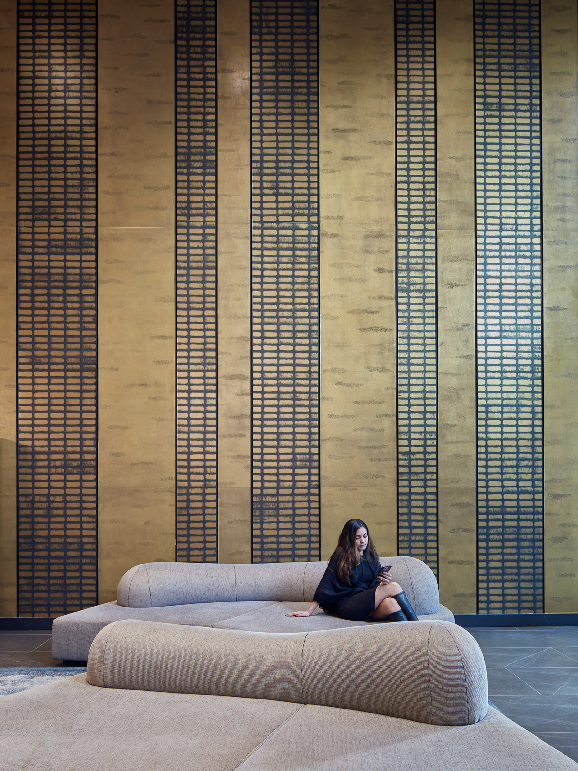 Sleek modern interior featuring a curved beige sofa centered on a grey area rug, with a wall behind adorned in vertical golden textured panels bisected by dark linear elements, creating a striking contrast. A woman sits casually on the sofa, engaging the space with a relaxed presence.