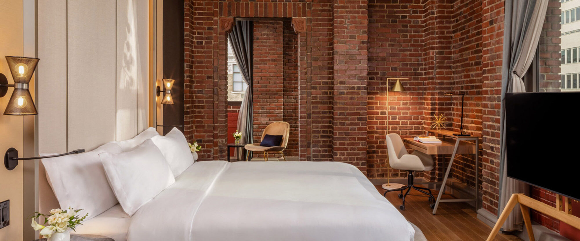 Elegantly appointed urban loft bedroom with exposed red brick walls and arched window, featuring a plush queen-sized bed, modernist lighting fixture above, and minimalist workspace to the side, blending industrial architecture with contemporary comfort.