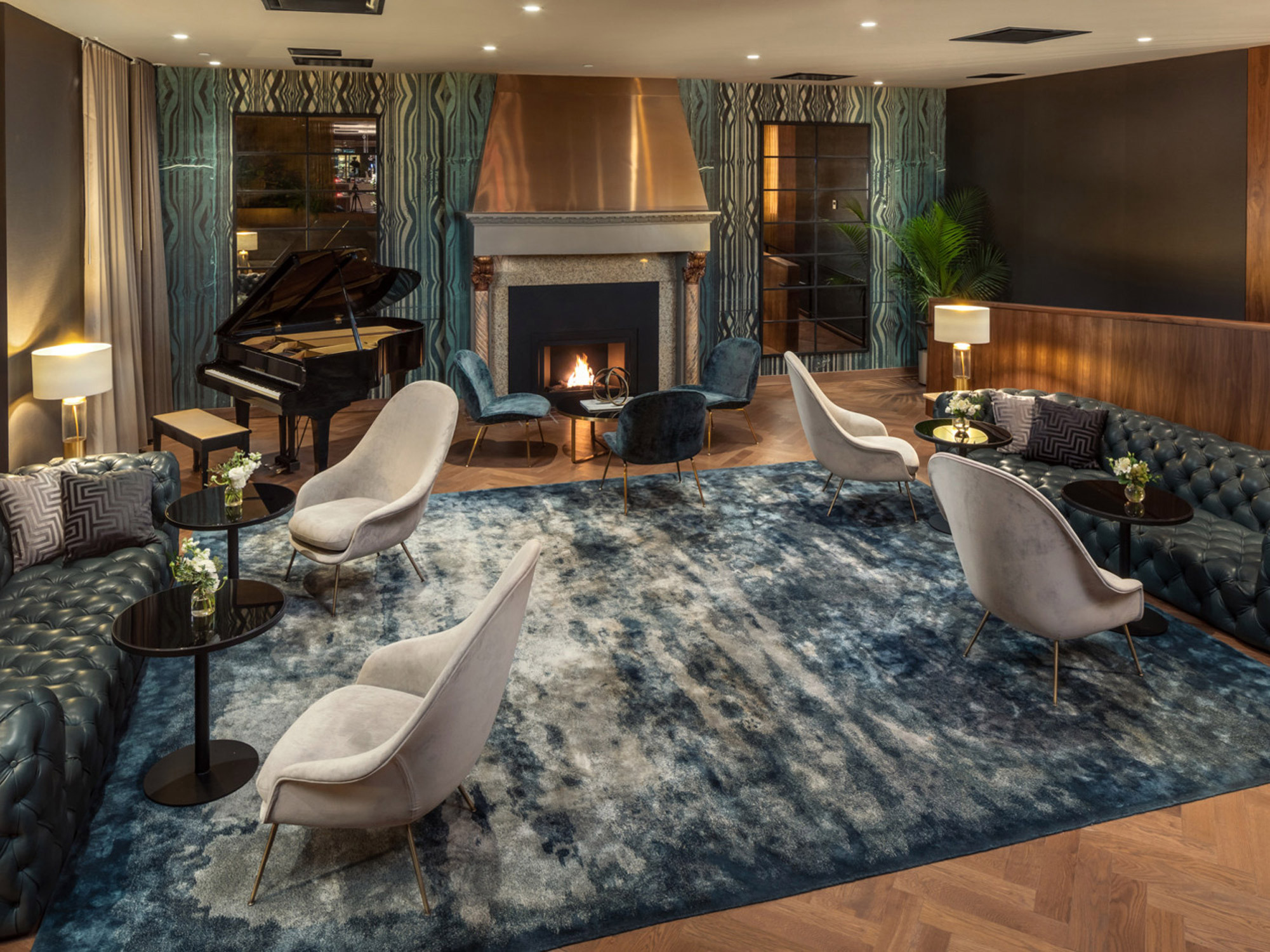 Elegant lounge area showcases a blend of textures, with a plush blue area rug anchoring sleek, ivory armchairs and a tufted leather sofa. A fireplace with a classic mantel serves as the focal point, flanked by rich teal curtains and a grand piano, enhancing the sophisticated ambiance.