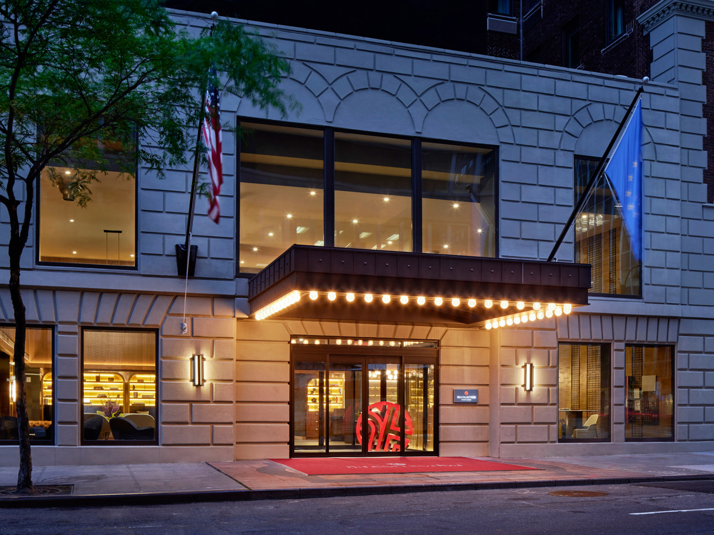 Exterior of a modern boutique hotel at dusk, showcasing an illuminated entryway with a red carpet, underlit awning, and a distinctive red sculpture visible through the glass doors.