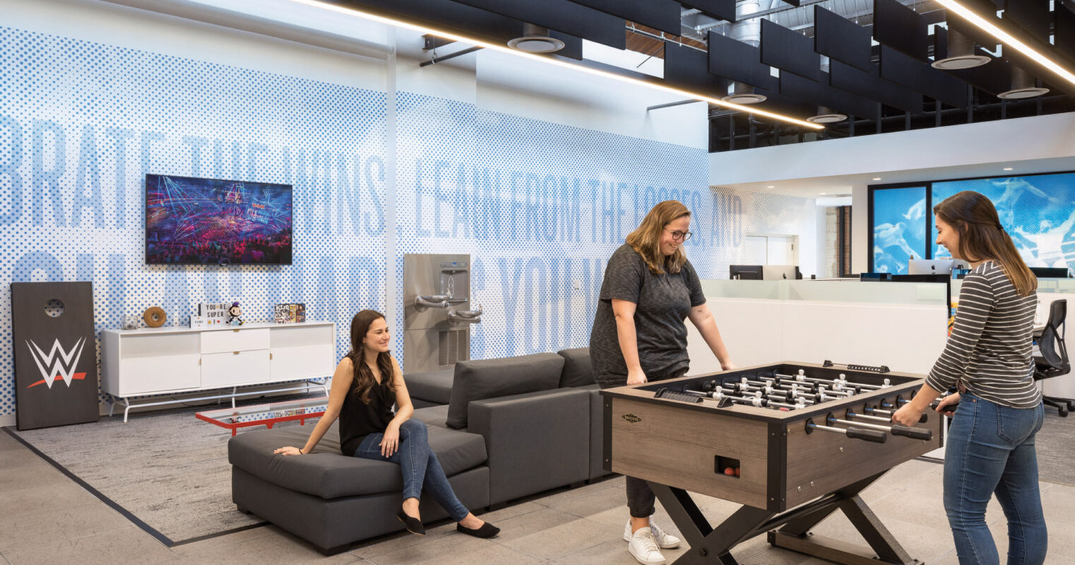 Modern office lounge with relaxed seating, a foosball table, and vibrant blue graphic wall featuring WWE branding. The space is designed for recreation, collaboration, and emphasizes the company's dynamic culture.