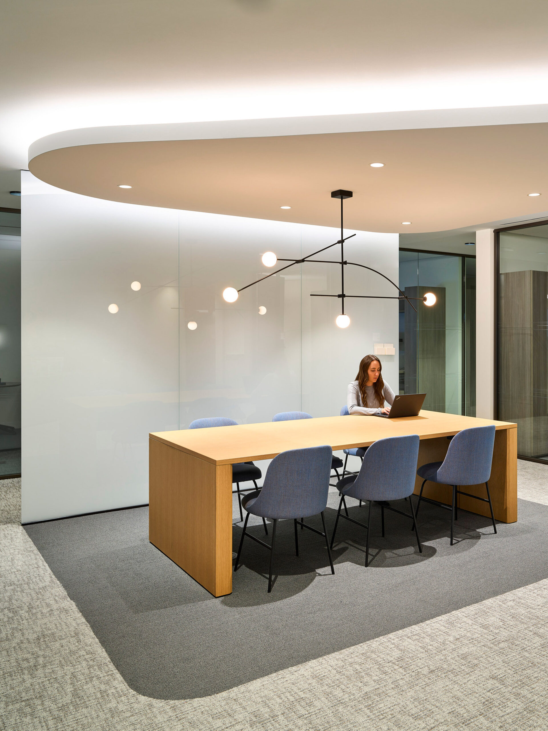 Modern office meeting space highlighted by an organic-shaped, floating ceiling panel and sleek pendant lighting. A central wooden table surrounded by blue upholstered chairs occupies the room, with a solitary professional working on a laptop, enhancing a focused yet collaborative ambiance.