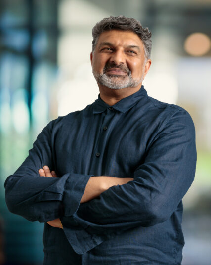 The photograph features a man with salt-and-pepper hair and a beard, giving a slight, confident smile, wearing a dark collared shirt with the sleeves rolled up to his elbows. He stands with his arms crossed in front of him against a light and airy office backdrop with out-of-focus lights, suggesting a professional environment.