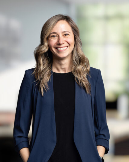 The image features a woman with long, wavy hair and a warm smile, wearing a black top and a navy blue blazer. The light and soft-focus background suggest she is in a modern and bright office setting.