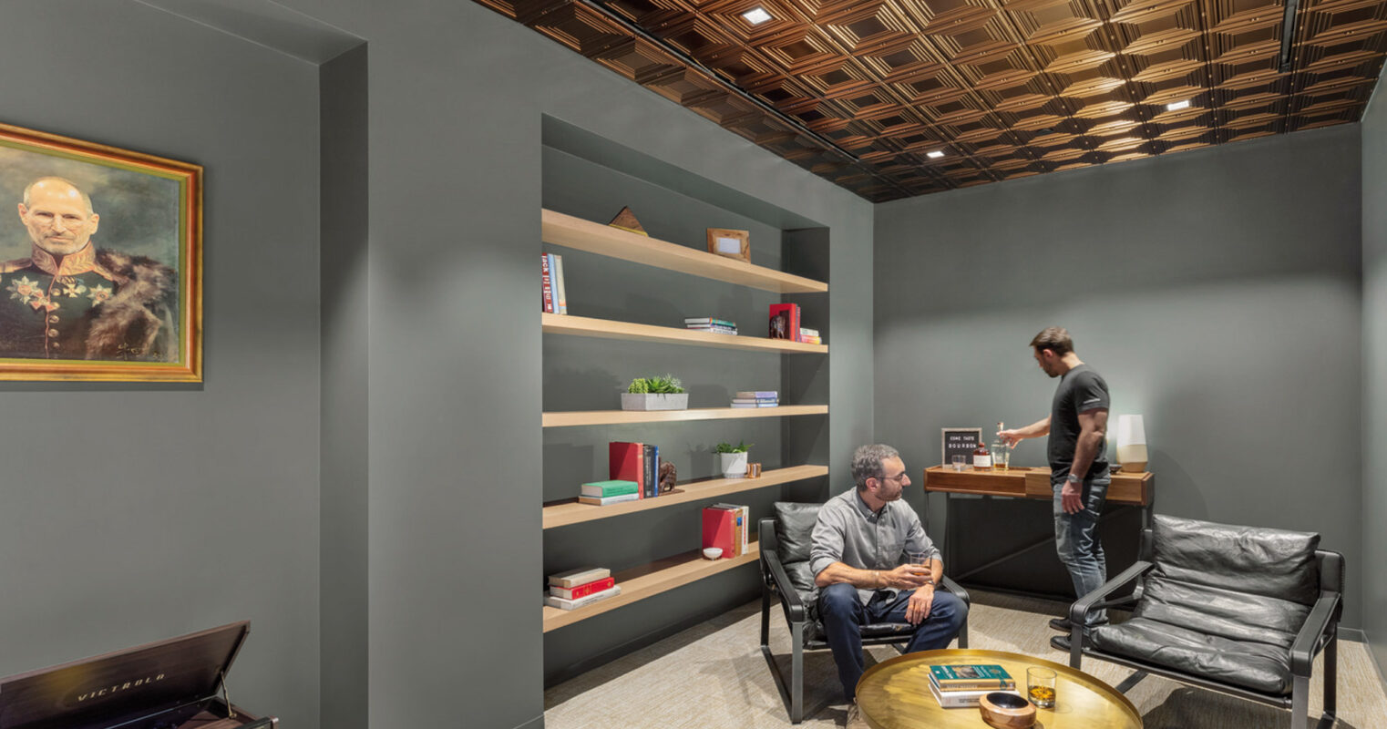 Modern office space with two individuals features charcoal-gray walls contrasting with a geometric copper ceiling. A built-in bookshelf, black leather chairs, and a central brass coffee table emphasize a sophisticated, contemporary aesthetic. A classic portrait adds a traditional touch to the room's sleek design.