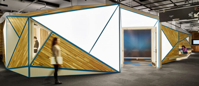 Modern office space featuring angular meeting pods with wooden slats and teal accents. Sleek, exposed ceiling fixtures add an industrial touch above the dynamic workspace layout.
