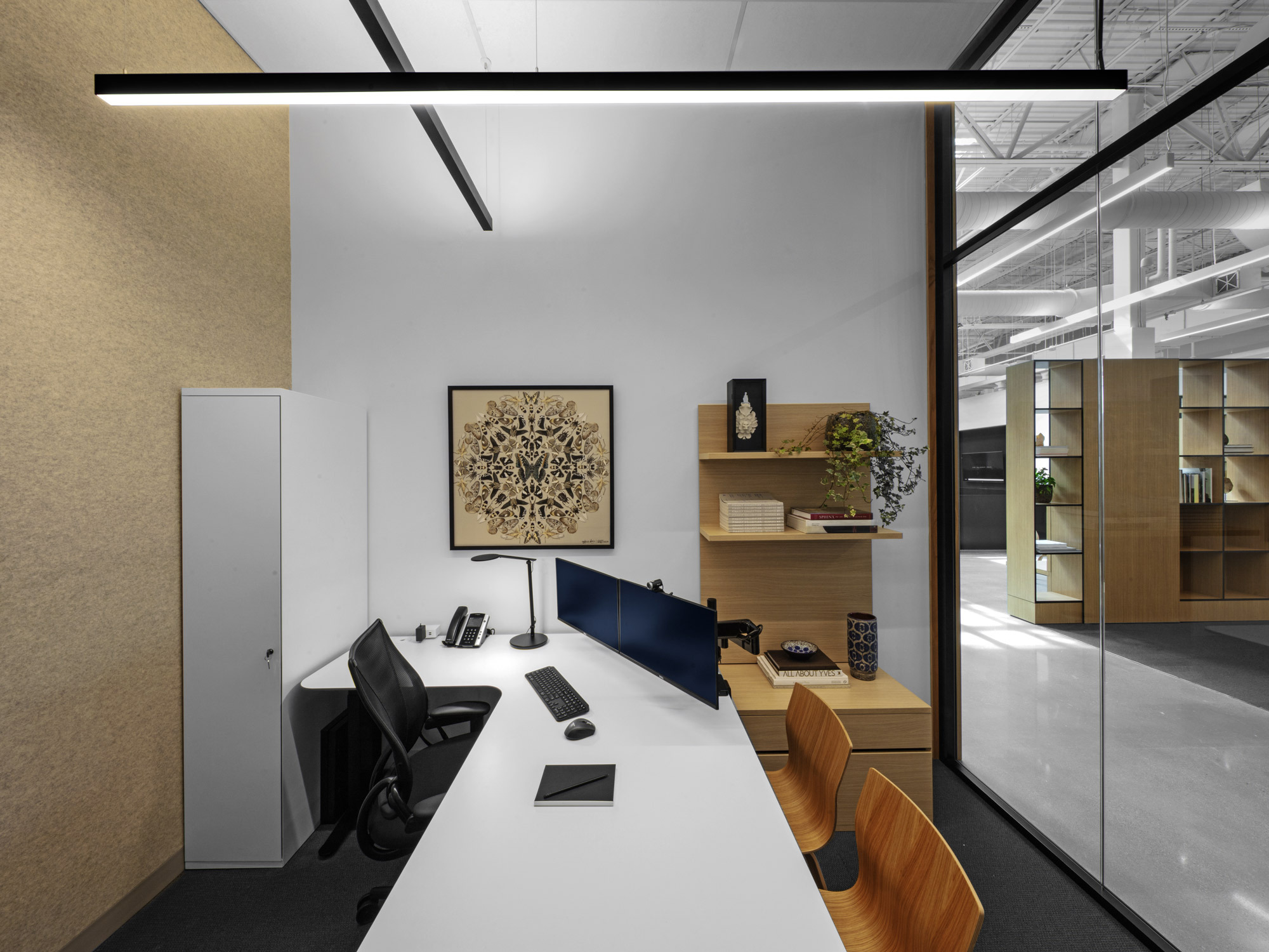 Minimalist office space boasting a sleek white desk with a desktop setup, ergonomic black chair, and decorative plants juxtaposed against a monochromatic color scheme, accentuated by geometric wall art and framed by linear suspended lighting. The glass partition reflects an open-plan environment beyond.