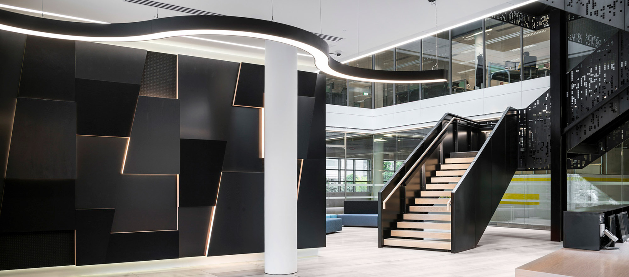 Modern office lobby featuring a sweeping white staircase with wooden steps, flanked by geometrically patterned black wall panels and illuminated by an undulating strip of overhead lighting. Clear glass balustrades outline mezzanine levels, illuminating the space with natural light.
