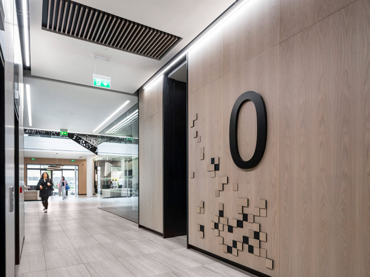 Modern corporate hallway showcasing natural wood panels contrasted by black accents, with unique geometric wall artwork and recessed linear lighting fixtures enhancing the streamlined design.
