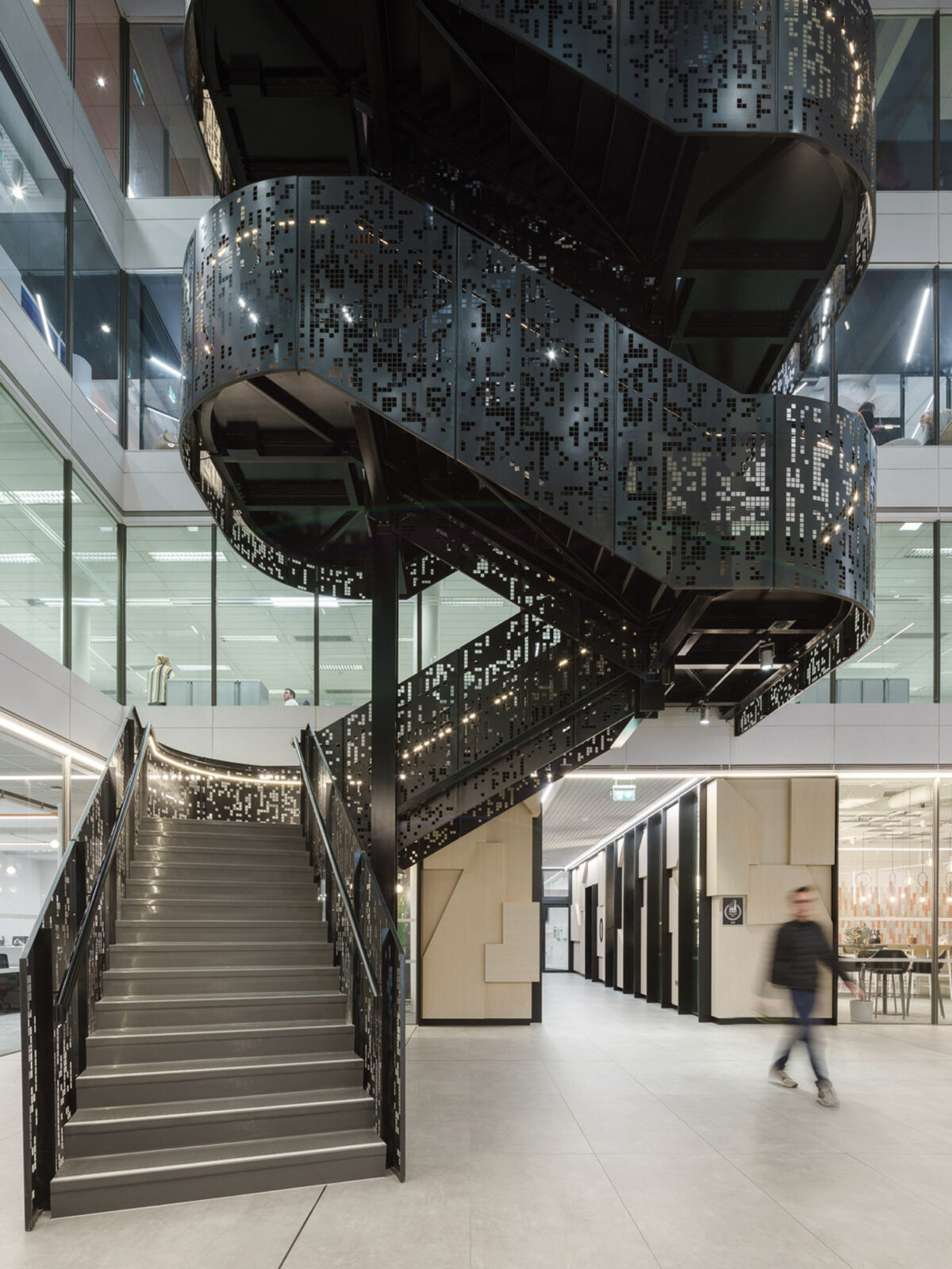 A modern atrium featuring a striking black spiral staircase with intricate geometric cutouts, creating a play of light and shadow. The surrounding minimalistic design accentuates the staircase's sculptural form.