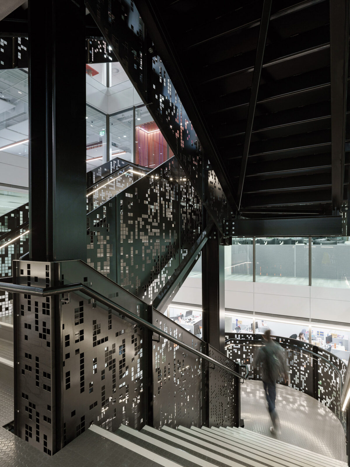 Modern staircase featuring perforated metal panels creating an urban geometric pattern. The ambient lighting accentuates the intricate shadows and highlights, contributing to the dynamic and contemporary aesthetic of the architectural space.