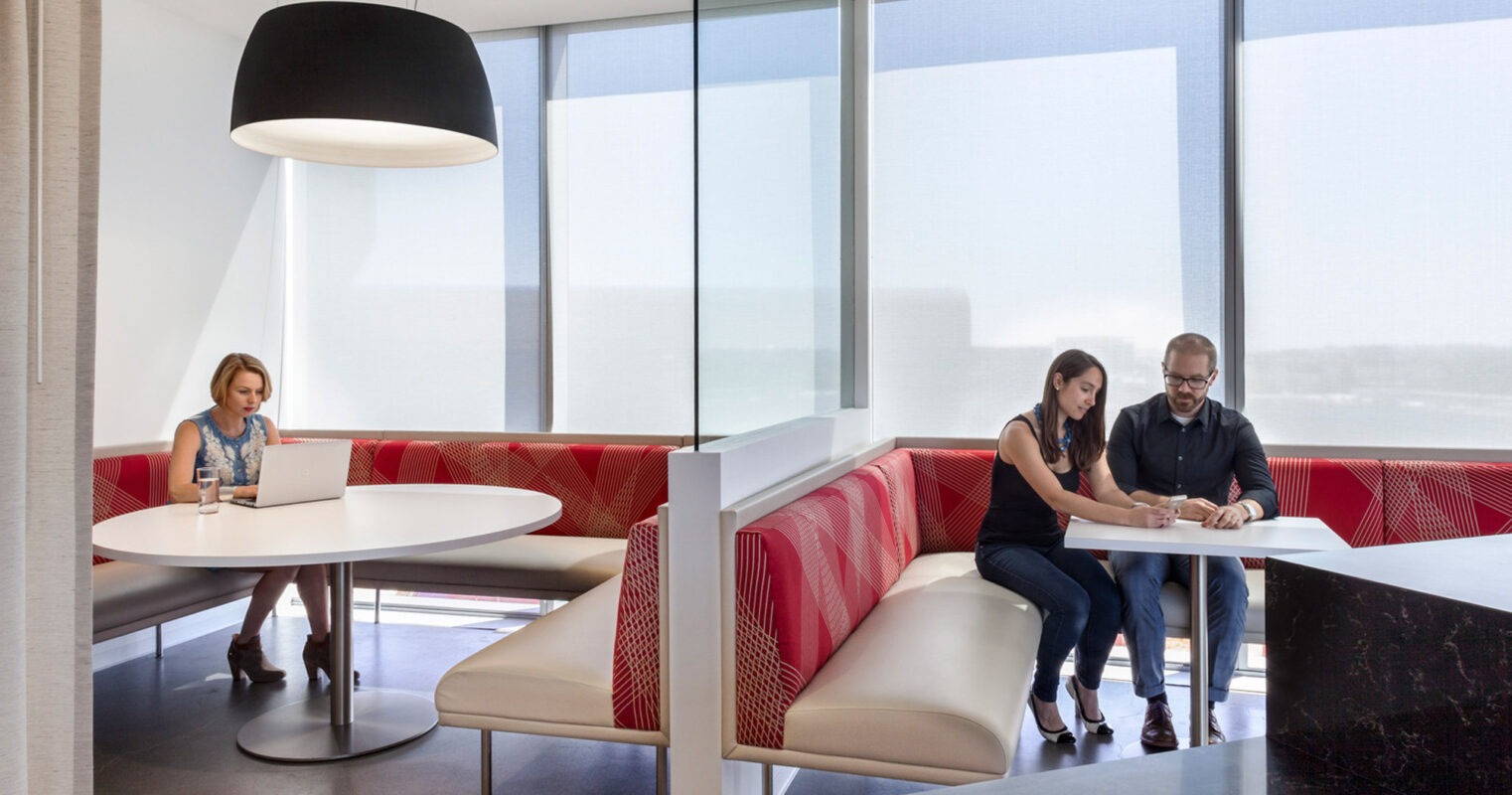 Modern office breakroom featuring a sleek white communal table, red geometric-patterned banquette seating, and an oversized white pendant light. Roller blinds diffuse sunlight, while employees engage in conversation and work on laptops, illustrating a dynamic, productive space with a striking color palette.