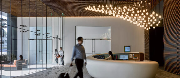 Sleek, modern office lobby featuring a curved white reception desk, accompanied by an array of hanging spherical lights. The ceiling showcases a dynamic grid of warm, wooden slats, enhancing the room's depth and texture. A blurred person in motion conveys the space's functionality.