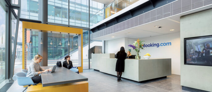 Modern office lobby with high ceilings and full-height windows, featuring a sleek white reception desk with a bold yellow accent panel. Two people work behind the counter, while others sit at a minimalist yellow bench, using laptops. The space is illuminated by abundant natural light.