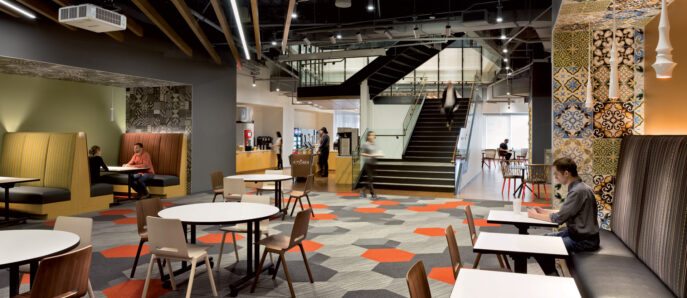 Modern office break room featuring eclectic design with exposed ceiling, patterned area rug, and mixed seating options including communal tables and booth-style benches. A staircase with metal railings ascends in the background, complemented by warm lighting and a decorative ceiling panel.