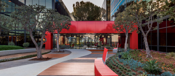 Landscape architecture integrates with modern corporate building design featuring a vibrant red awning, matching linear benches, and a meandering pathway amidst drought-tolerant plantings.