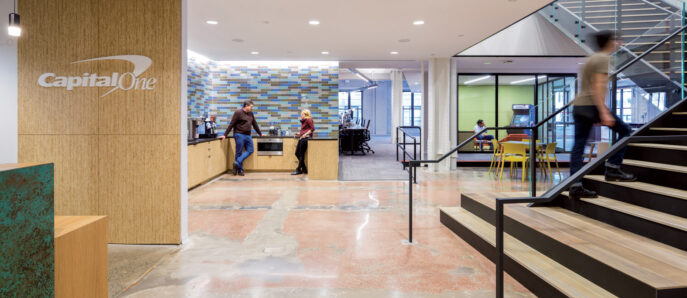 Spacious commercial lobby with mix of natural and industrial elements, featuring a terrazzo floor, wood-paneled reception desk with the Capital One logo, mosaic tile wall art, and a staircase leading to an upper level with occupants in motion, reflecting dynamic workplace design.