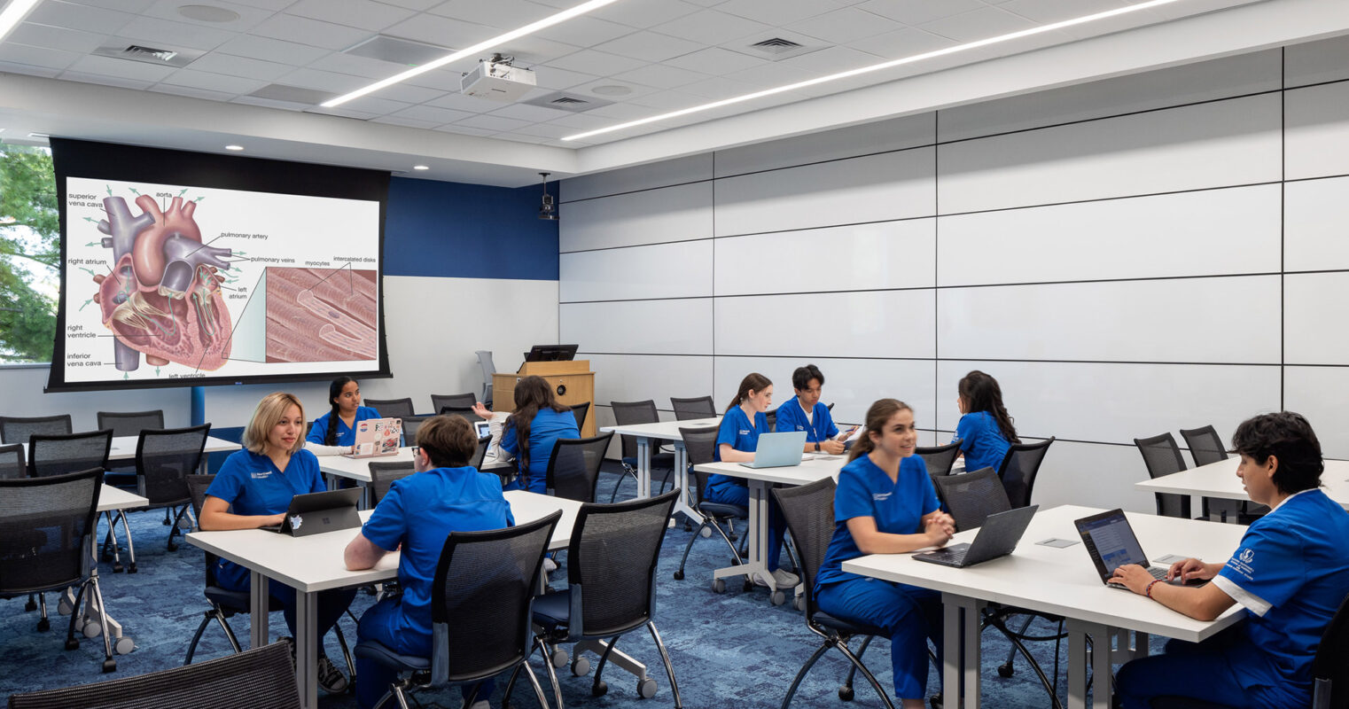 Spacious classroom featuring modern movable tables and chairs with a blue and white color scheme. The front wall displays a large projection screen, while ample natural light enhances the learning environment.