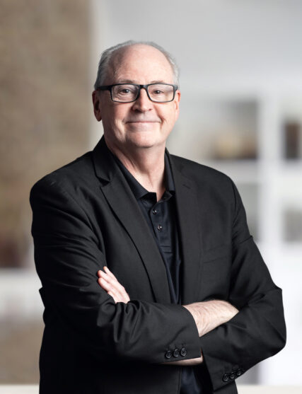 The image features a man with glasses and grey hair, dressed in a black shirt and blazer, with his arms crossed in a confident stance. The soft, neutral background suggests an indoor setting with a professional atmosphere.