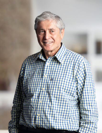 The photograph features an older man with silver hair and a friendly demeanor, wearing a blue and white checked shirt. He is posed against a soft-focused background that has architectural elements, indicating a professional environment.