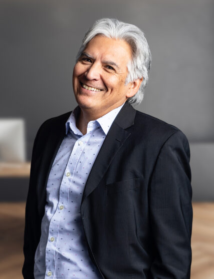 The image depicts a man with silver hair and a genuine smile, wearing a dark suit jacket over a light blue shirt with a subtle pattern. The neutral grey background is softly focused, highlighting the subject in a professional setting.