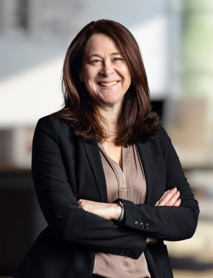 The image shows a woman with shoulder-length brown hair and a content smile, dressed in a professional black blazer and beige blouse. She stands with her arms crossed, conveying a confident and approachable stance against a softly blurred background suggestive of an office space.
