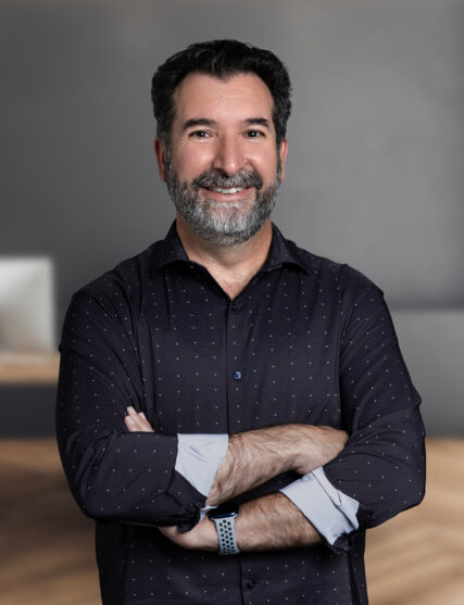 A portrait of a cheerful man with dark hair and a short beard, arms crossed and sporting a friendly smile. He is dressed in a dark button-down shirt with a subtle pattern, complemented by rolled-up cuffs, in a modern office setting.
