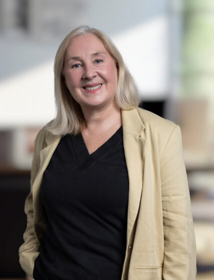A professional portrait of a woman with long, straight blonde hair, smiling gently at the camera. She is wearing a beige blazer over a black top, and her relaxed yet confident pose in an office setting suggests experience and authority.