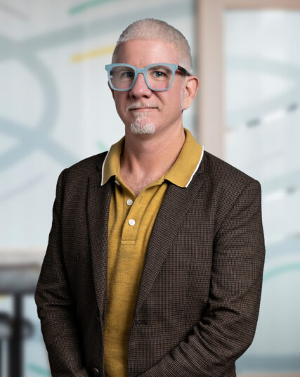 A professional portrait of a man with graying hair, a mustache, and beard standing confidently in an office environment. He's wearing blue-rimmed glasses, a mustard-colored shirt, and a brown checkered blazer, giving off a look of experienced leadership.