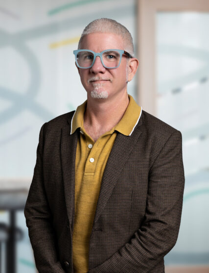 A professional portrait of a man with graying hair, a mustache, and beard standing confidently in an office environment. He's wearing blue-rimmed glasses, a mustard-colored shirt, and a brown checkered blazer, giving off a look of experienced leadership.
