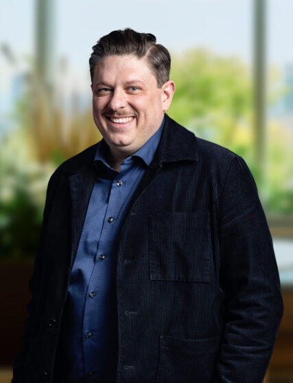 The image displays a man with a cheerful expression and neatly styled hair, dressed in a blue shirt and a dark textured coat. He stands with a slight lean forward, against a backdrop of blurred natural elements and daylight, giving the impression of a comfortable and inviting workspace.