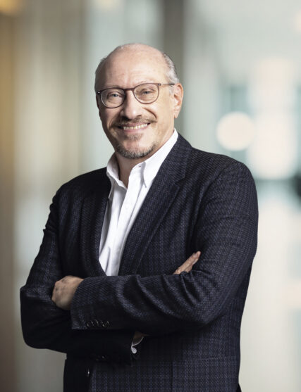 The image shows a man with a beard and glasses, sporting a confident smile, dressed in a dark textured blazer over a crisp white shirt. He stands with his arms folded, projecting a relaxed yet authoritative demeanor, against a softly blurred background typical of a professional office environment.