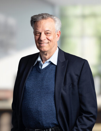 The image depicts a mature man with greying hair and a friendly smile, wearing a dark suit jacket over a blue sweater and a collared shirt. He stands with his arms slightly crossed, radiating confidence and professionalism against a blurred office background.