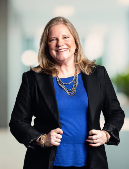 The image displays a woman with long blonde hair and a joyful smile, wearing a vibrant blue top, a black blazer, and gold jewelry. She stands with her hands on her hips in a confident pose, set against a blurred backdrop indicative of a bright and modern office space.