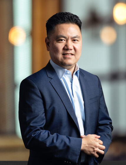 The image shows a man with a confident stance and a friendly expression, sporting short hair, dressed in a navy blue suit and light blue shirt. He stands with crossed arms, set against a warm, blurred backdrop that suggests an inviting and professional interior space.