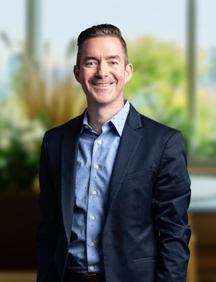 The image captures a man with a friendly smile, sporting a short haircut, wearing a dark suit jacket over a light blue, patterned shirt. He stands with his hands casually at his sides against a backdrop of soft, natural light and blurred greenery, suggesting an office setting with a view of the outdoors.