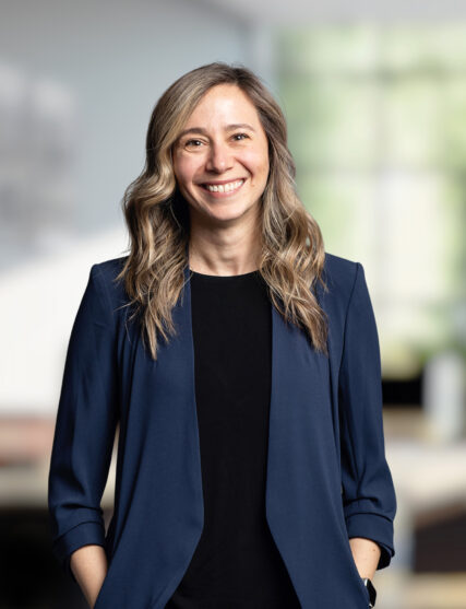 The image features a woman with long, wavy hair and a warm smile, wearing a black top and a navy blue blazer. The light and soft-focus background suggest she is in a modern and bright office setting.