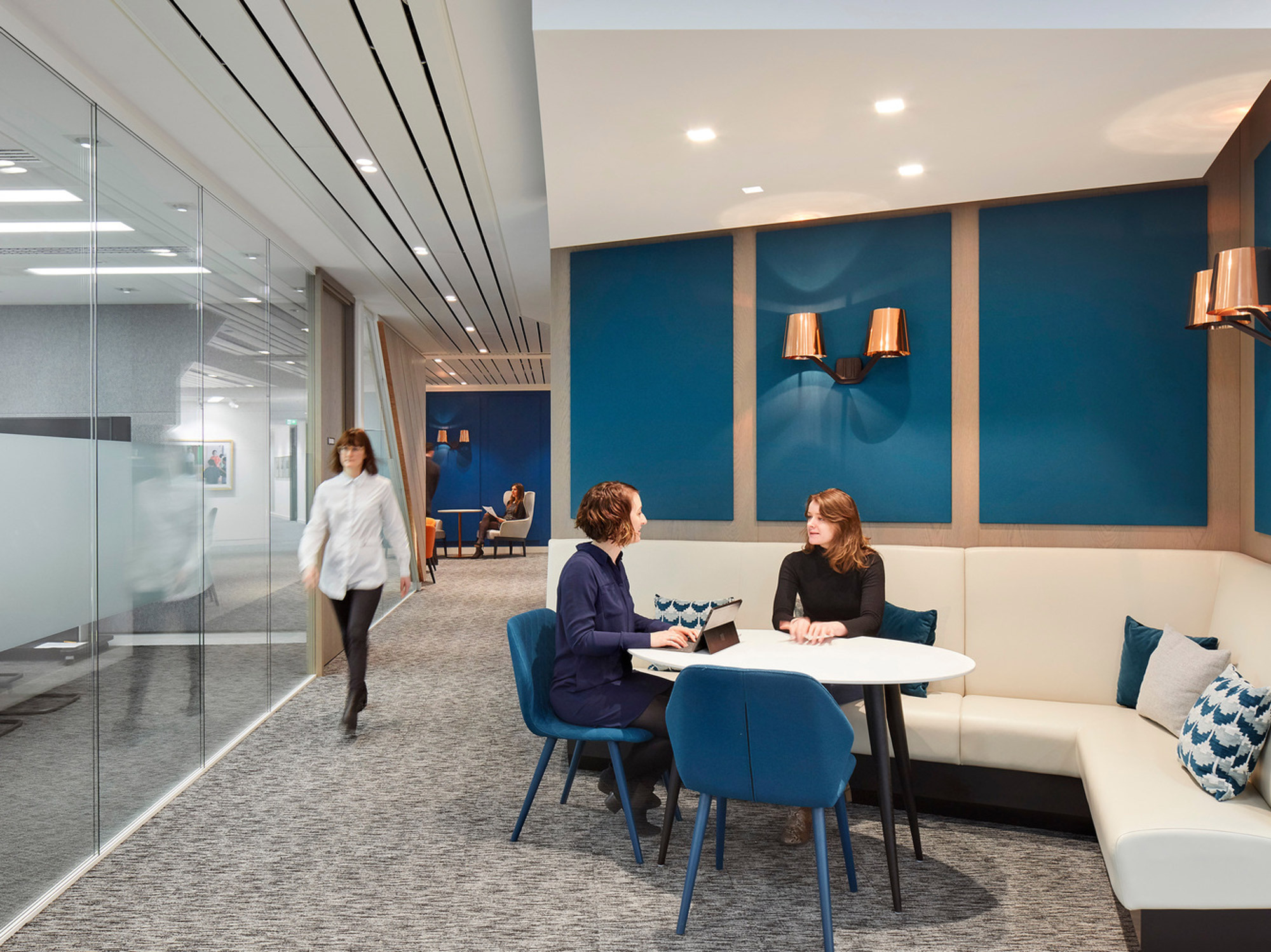 Modern office lounge showing a conversation area with a plush white couch, blue privacy panels, wood accents, and sleek wall-mounted sconces providing warm ambient lighting. A person strides through the hallway beyond the cozy seating arrangement.