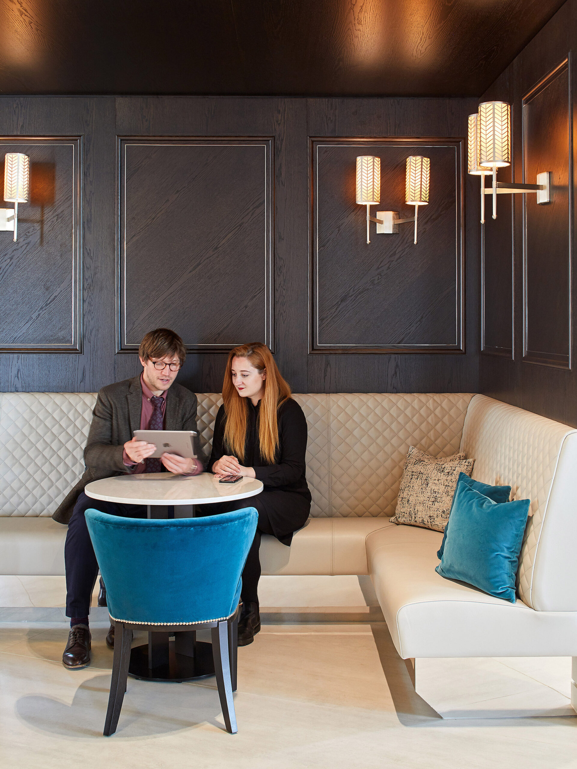 Two professionals confer in an elegantly designed meeting space with dark paneled walls and luxurious textures. A cream sofa with plush cushions, contemporary wall sconces, and a royal blue velvet chair underline the room’s sophisticated color palette and lighting strategy.