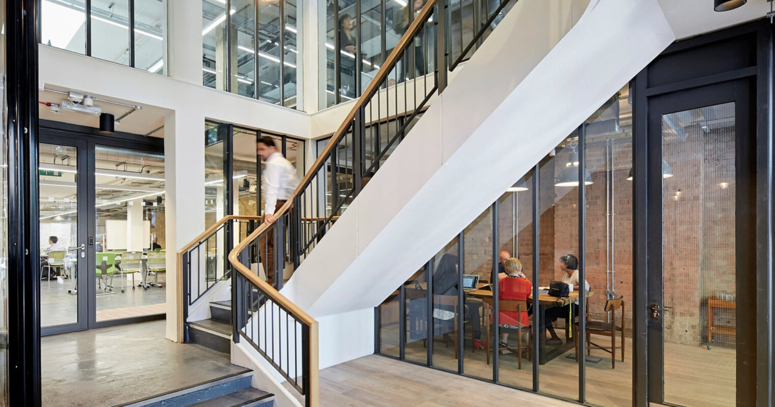 Modern office interior featuring a minimalist staircase with sleek metal railings, light wooden steps, and adjacent glass panels allowing visibility into collaborative workspace areas with exposed brick walls and contemporary furnishings. Natural light floods in from generous skylights above.