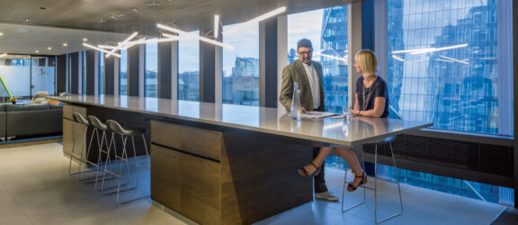 Modern office interior with two professionals engaged in a discussion at a sleek, wooden standing desk. Linear pendant lighting accents the urban skyline view through floor-to-ceiling windows, complementing the space's open and contemporary aesthetic.