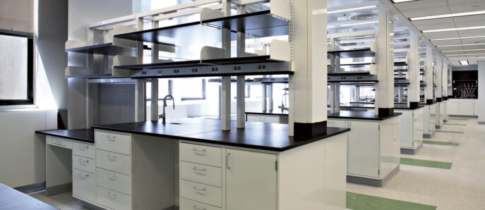 Modern laboratory interior featuring rows of sleek, white workstation benches with integrated shelving. Overhead, exposed ductwork contrasts with linear lighting fixtures, and green floor markings guide traffic flow, reflecting a functional yet aesthetically clean design.
