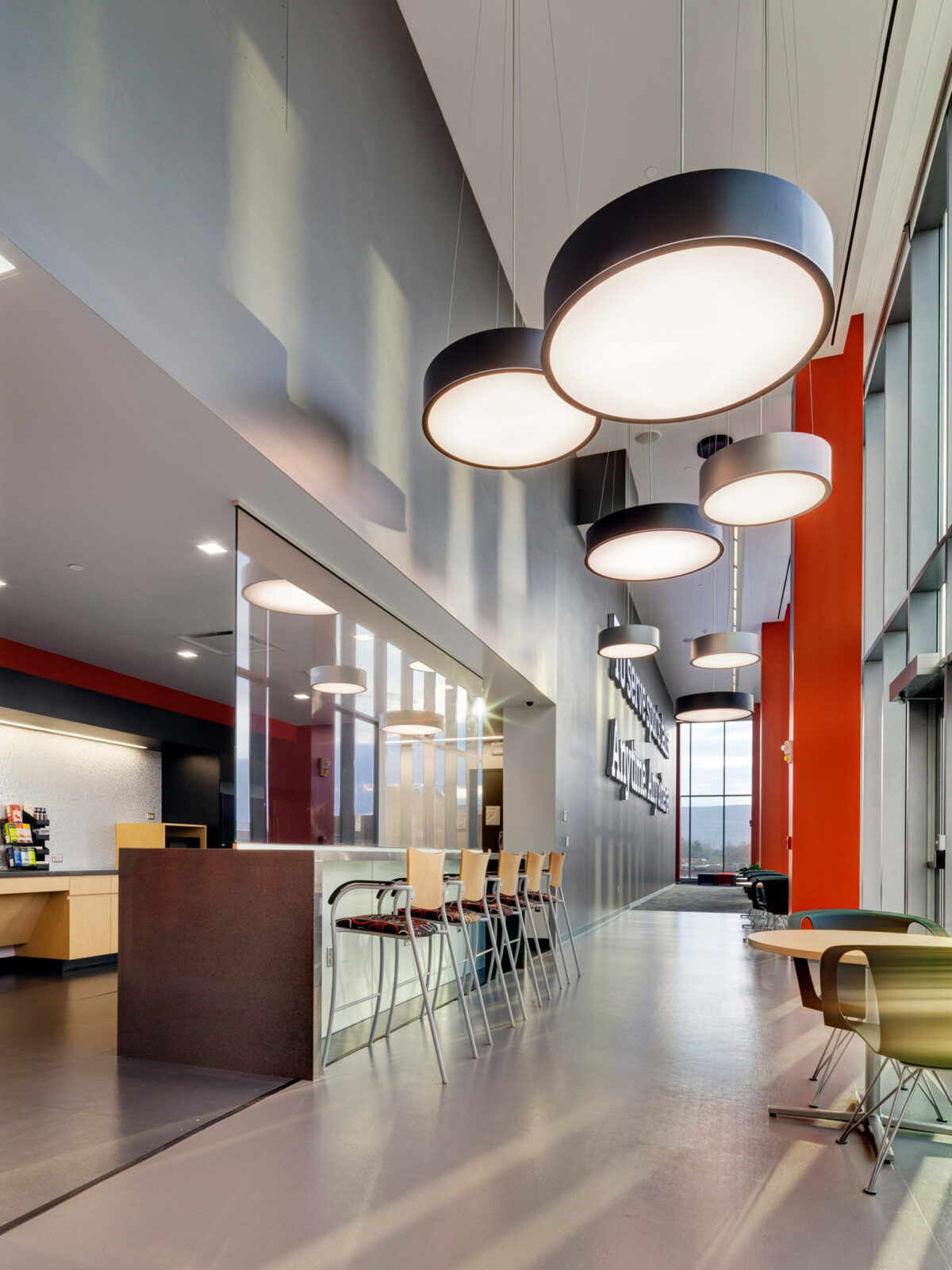 Spacious modern lobby with sleek gray flooring. Large circular pendant lights hang above, complementing the vibrant red accent walls. Minimalist furniture is strategically placed beside expansive glass partitions, contributing to a contemporary aesthetic.