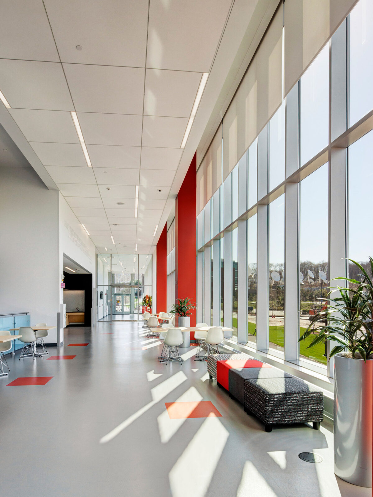 Bright, modern office lounge with floor-to-ceiling windows, offering ample natural light. White designer chairs and black ottoman complement the minimalist aesthetic. Red accent wall adds a pop of color amidst a neutral palette, enhancing the space's contemporary feel.