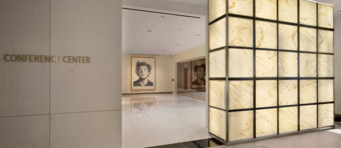 Elegantly lit translucent stone paneling flanks the entrance to a modern conference center, contrasting with a minimalistic matte wall bearing the center's signage. The warm glow adds ambience, harmonizing with the reflective flooring while highlighting adjacent artwork.