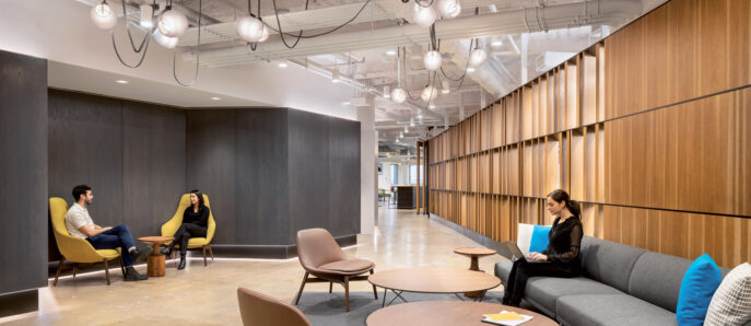 Spacious office lounge area featuring a blend of natural wood paneling and exposed concrete ceiling. Mid-century modern furniture with rounded tables and sleek chairs offer informal seating arrangements. Overhead, distinctive linear pendant lighting complements the contemporary workspace ambiance.