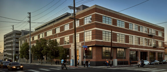 Twilight view of a renovated industrial building converted into modern office space, showcasing red brick exterior walls, large symmetrical windows, and a prominent corner entrance. The design combines historical architecture with contemporary urban style.