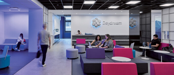 Modern office lounge featuring blue and magenta furniture with clean lines, accented by dynamic geometric carpet patterns. A boldly branded wall provides a backdrop to collaborative seating areas, emphasizing open-concept design and facilitating informal teamwork.