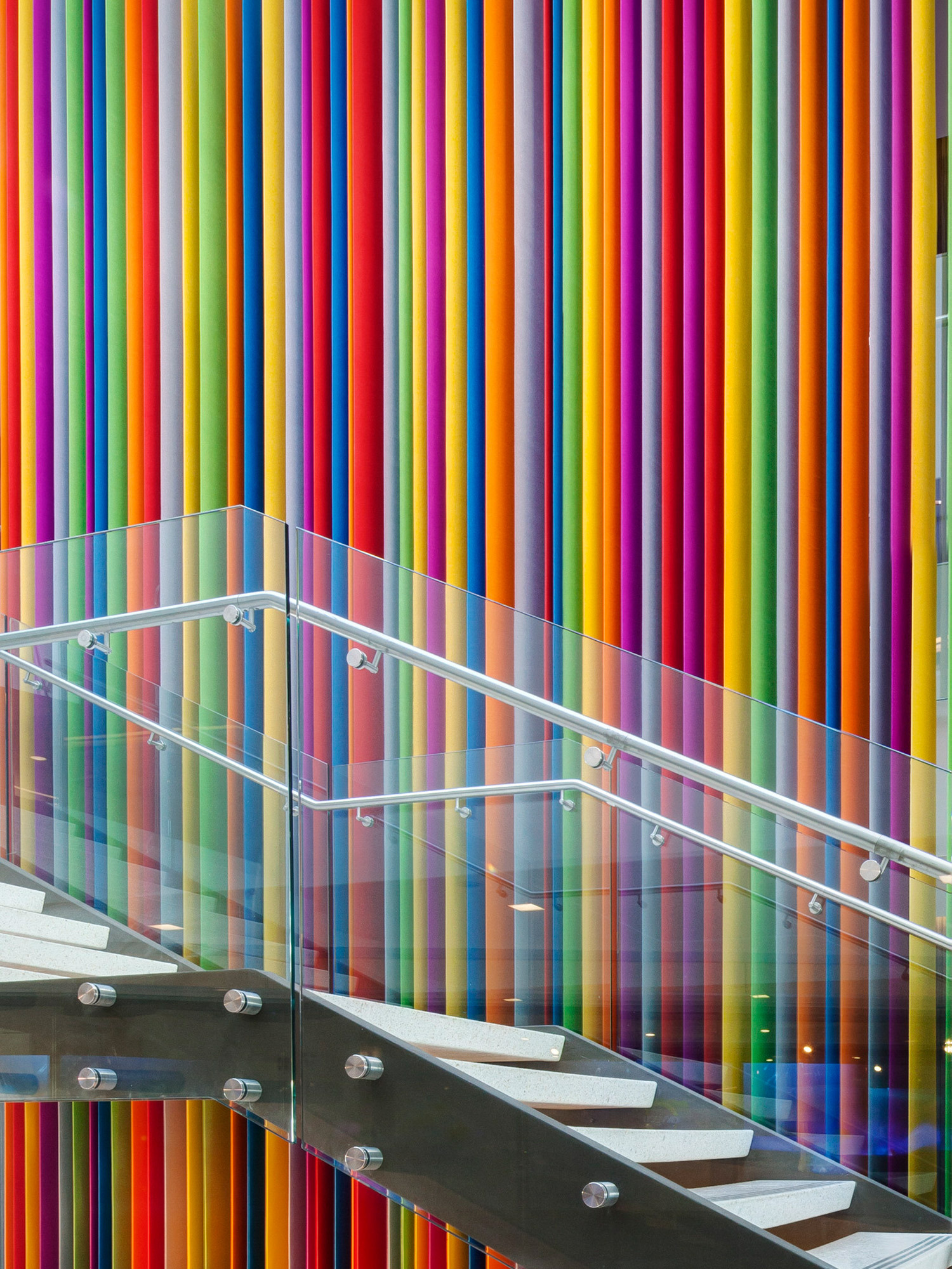 Modern staircase alongside a vibrant, multicolored vertical striped wall feature. Glass balustrades provide a minimalist contrast to the playful, rainbow-colored backdrop, illustrating a bold use of color in contemporary interior design.