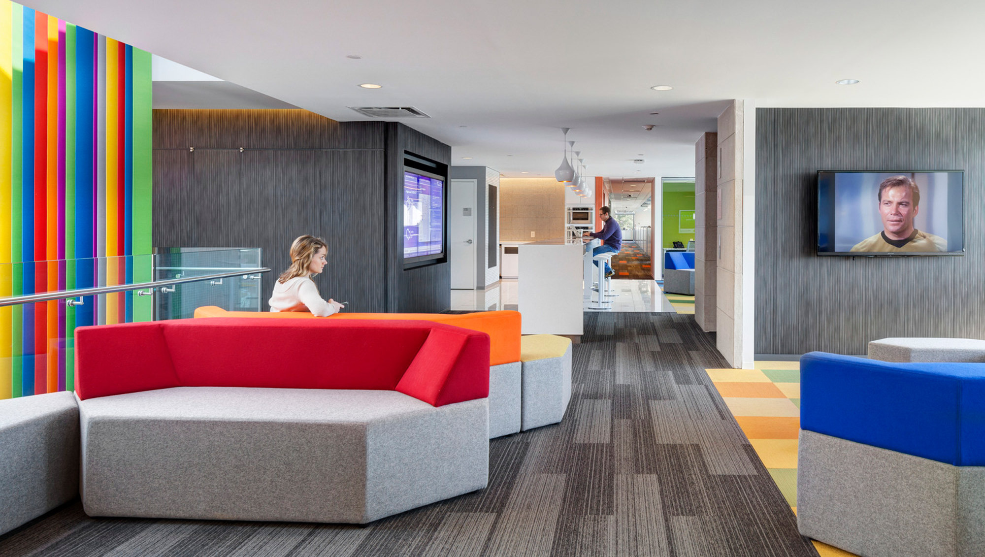 Modern open-plan office lounge featuring vibrant color-blocked accents, a red-orange modular sofa, and gray upholstered seating, set against subtle wood paneling and a lively carpet with geometric patterns. The space is casually divided with a partial floating wall hosting a television screen.